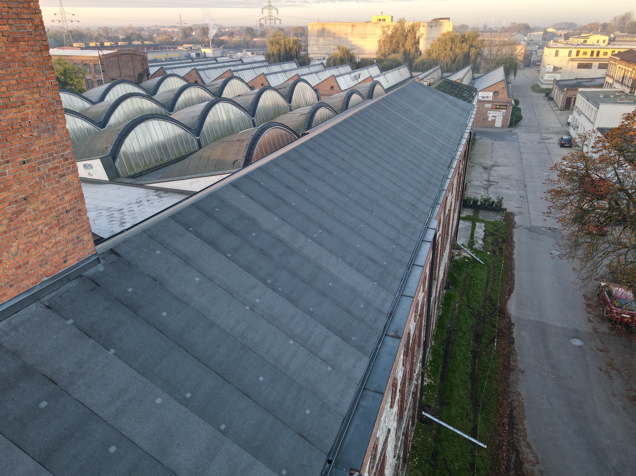 Rozległy, płaski dach przemysłowy pokryty papą, widok z góry na tle zabudowań fabrycznych i ulicy z zaparkowanym samochodem.