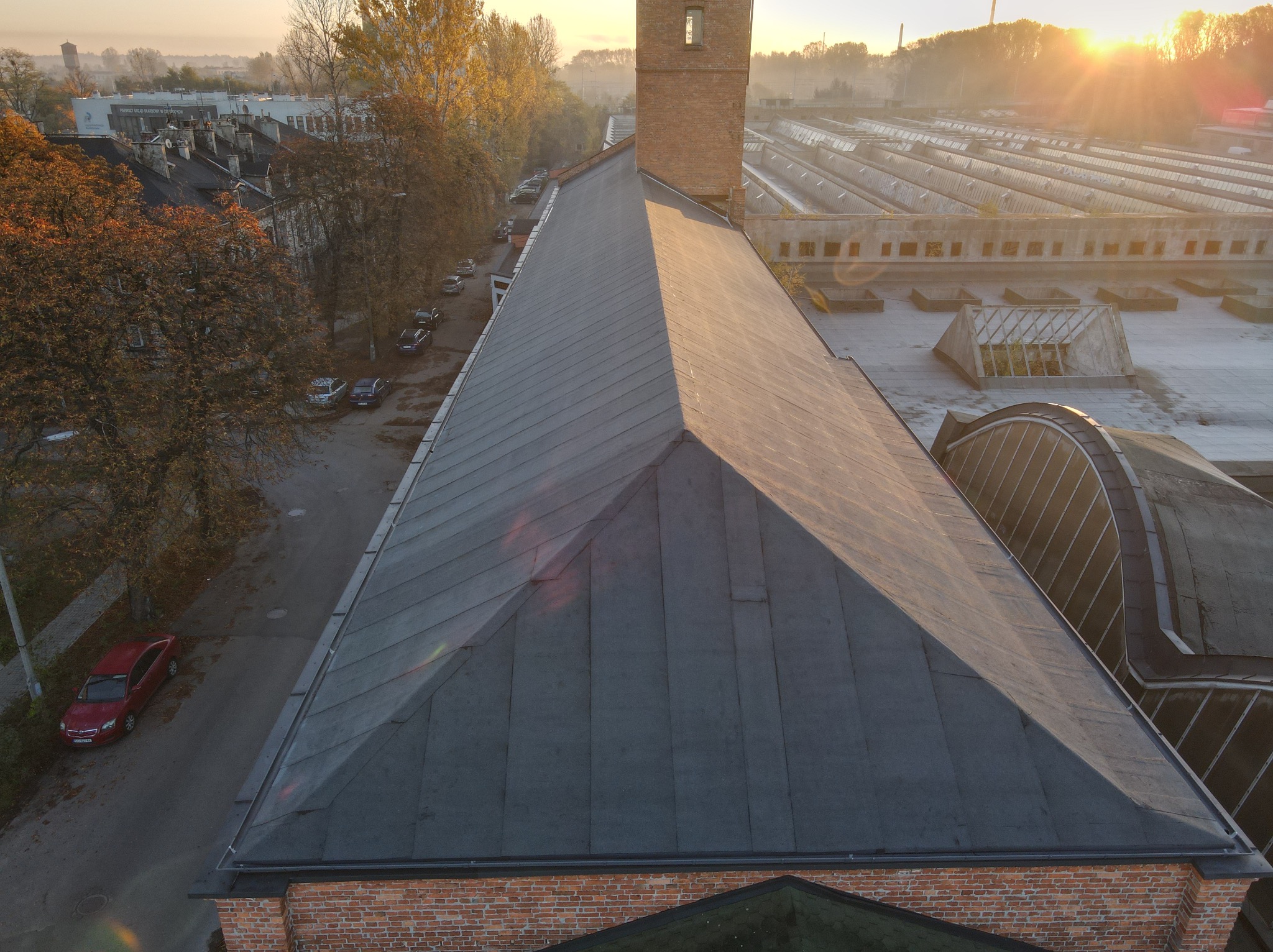 Widok z góry na duży, płaski dach pokryty papą, z ceglaną wieżą po lewej stronie i przemysłowymi halami w tle, oświetlony wschodzącym słońcem.