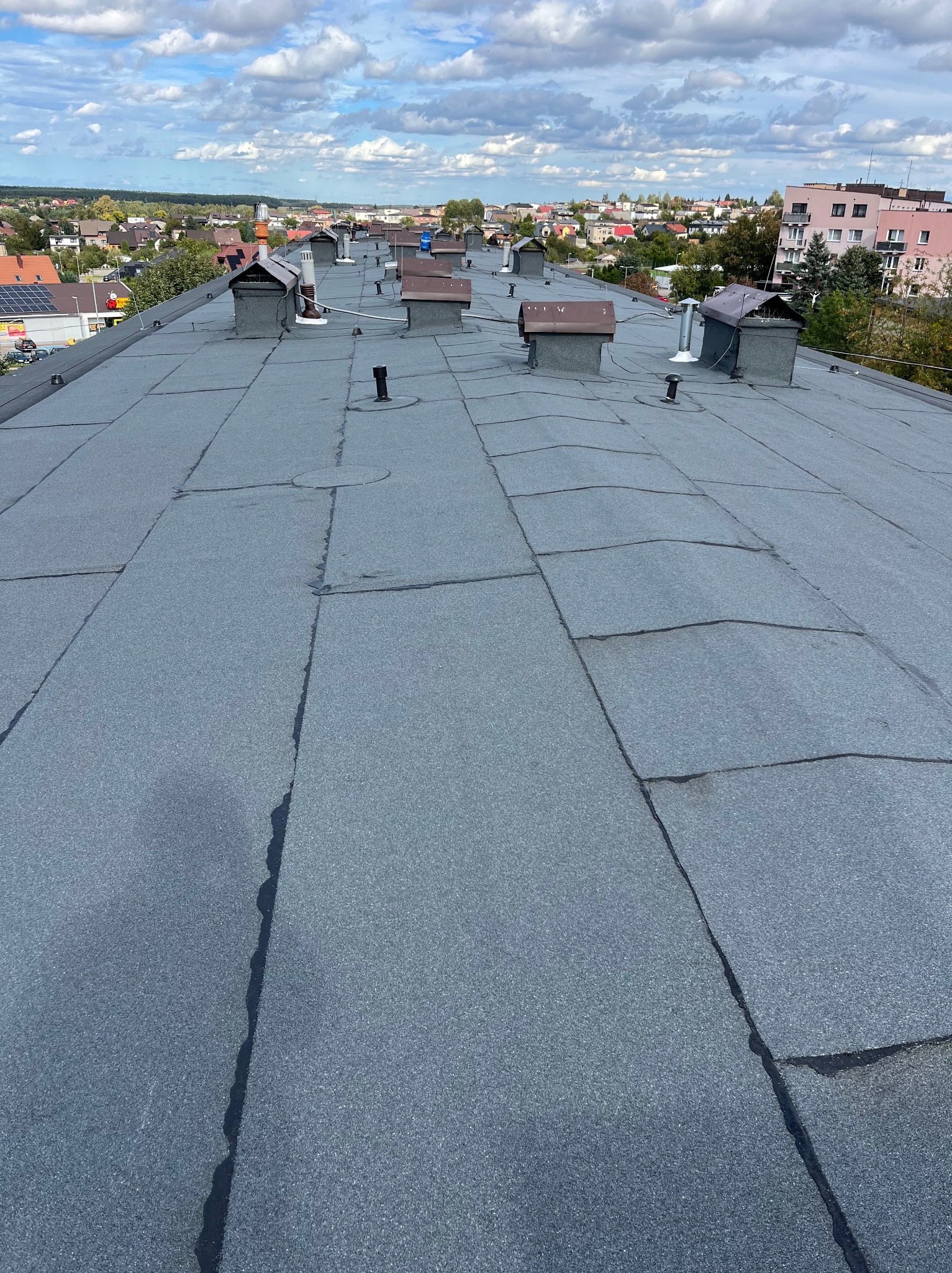 Rozległy, płaski dach pokryty papą termozgrzewalną z licznymi kominami wentylacyjnymi na tle zabudowy miejskiej i błękitnego nieba z obłokami. Widoczne łączenia papy oraz drobne uszkodzenia.