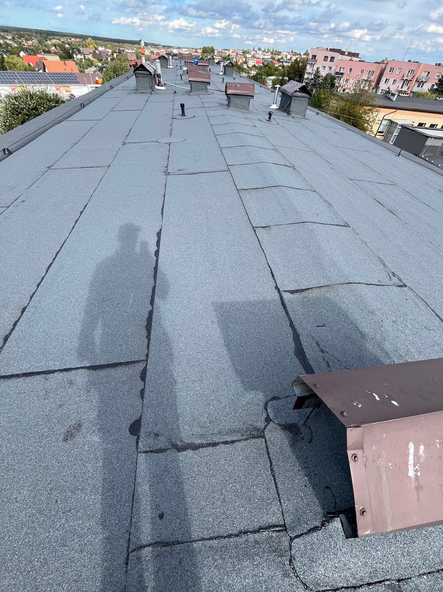 Szary dach z papy w Częstochowie z widocznym cieniem osoby wykonującej prace, komin wentylacyjny i obróbka blacharska komina.