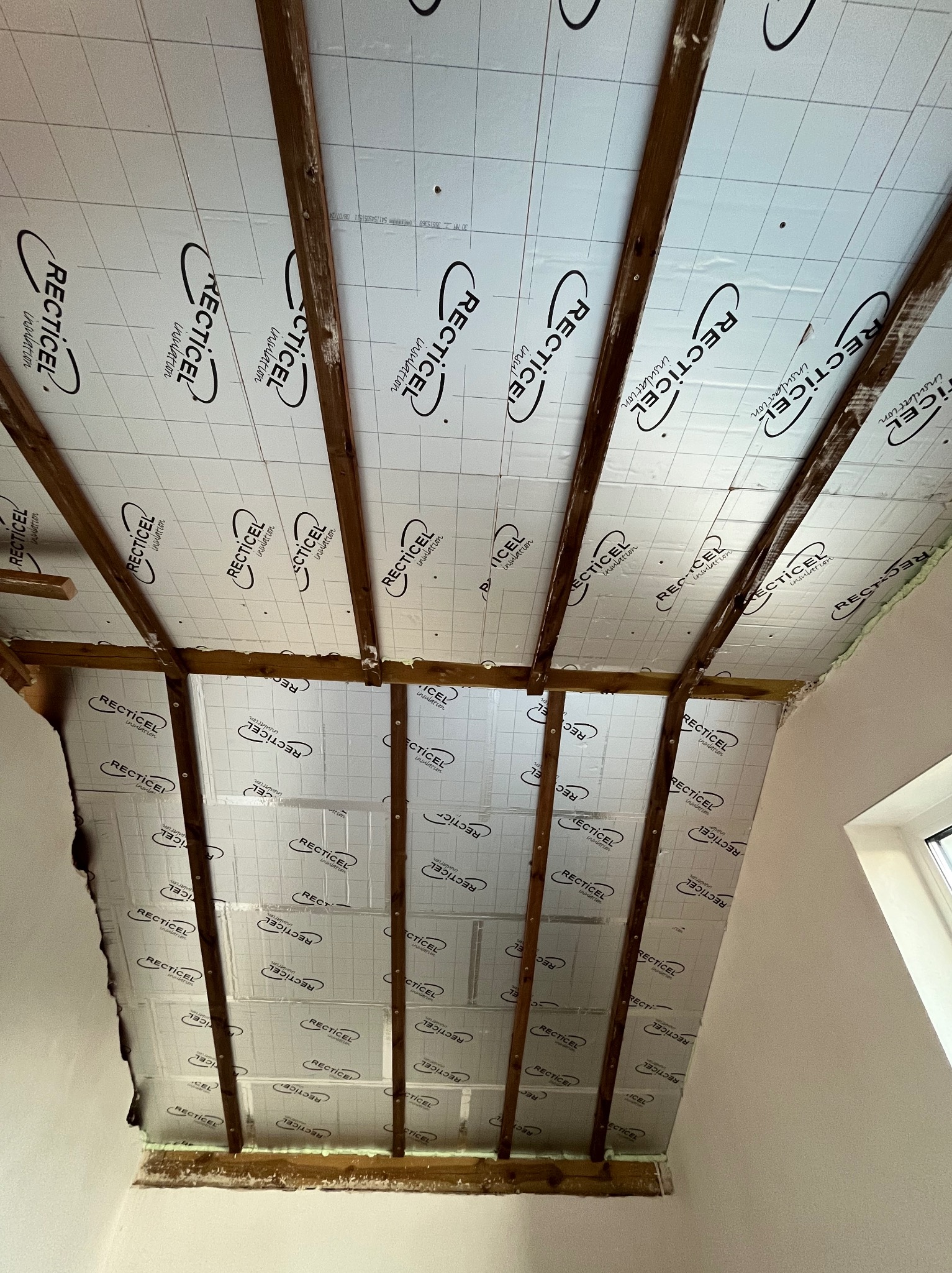 Wnętrze poddasza w trakcie prac: drewniana więźba dachowa z zamontowaną izolacją z folią z nadrukowanym logo producenta, widoczne fragmenty ściany i okna.