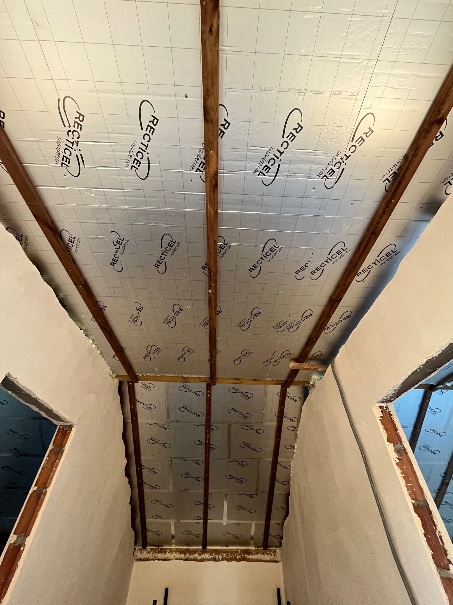 Wnętrze poddasza w trakcie ocieplania: widoczne drewniane elementy więźby dachowej oraz srebrna folia izolacyjna z nadrukowanym logo Recticel, surowe ściany z widocznymi fragmentami cegieł.