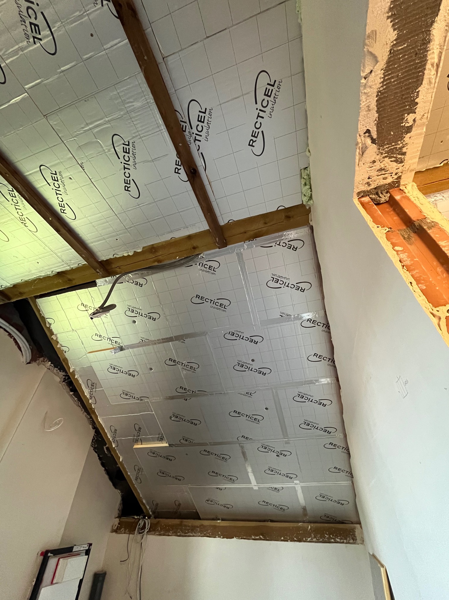 Montaż izolacji dachu z widocznymi drewnianymi belkami i folią izolacyjną z nadrukiem, fragment ściany z widocznymi warstwami wykończeniowymi.