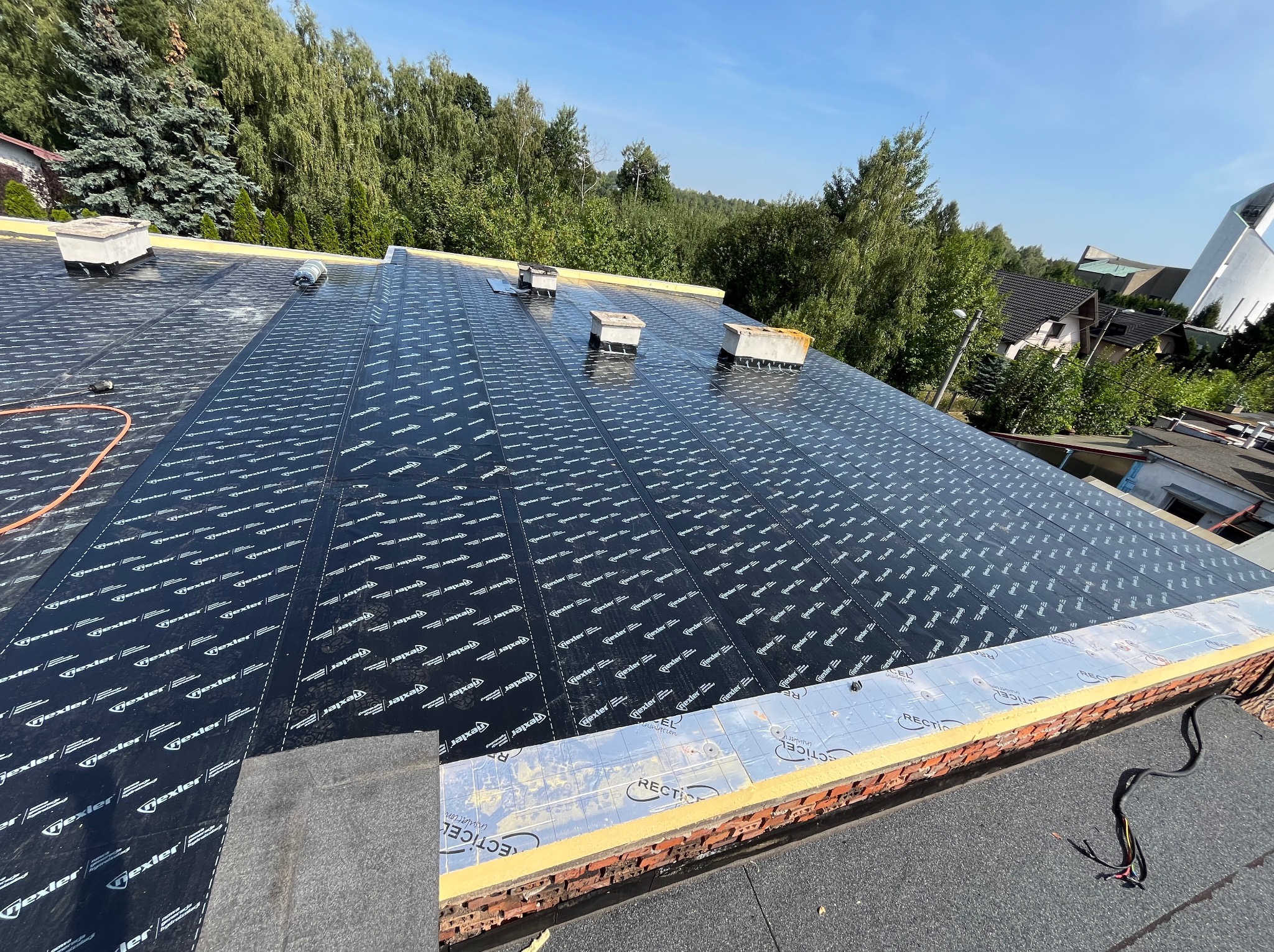 Dach pokryty czarną membraną z nadrukiem, widoczne kominy wentylacyjne i fragment muru z izolacją termiczną, na tle zieleni i błękitnego nieba.