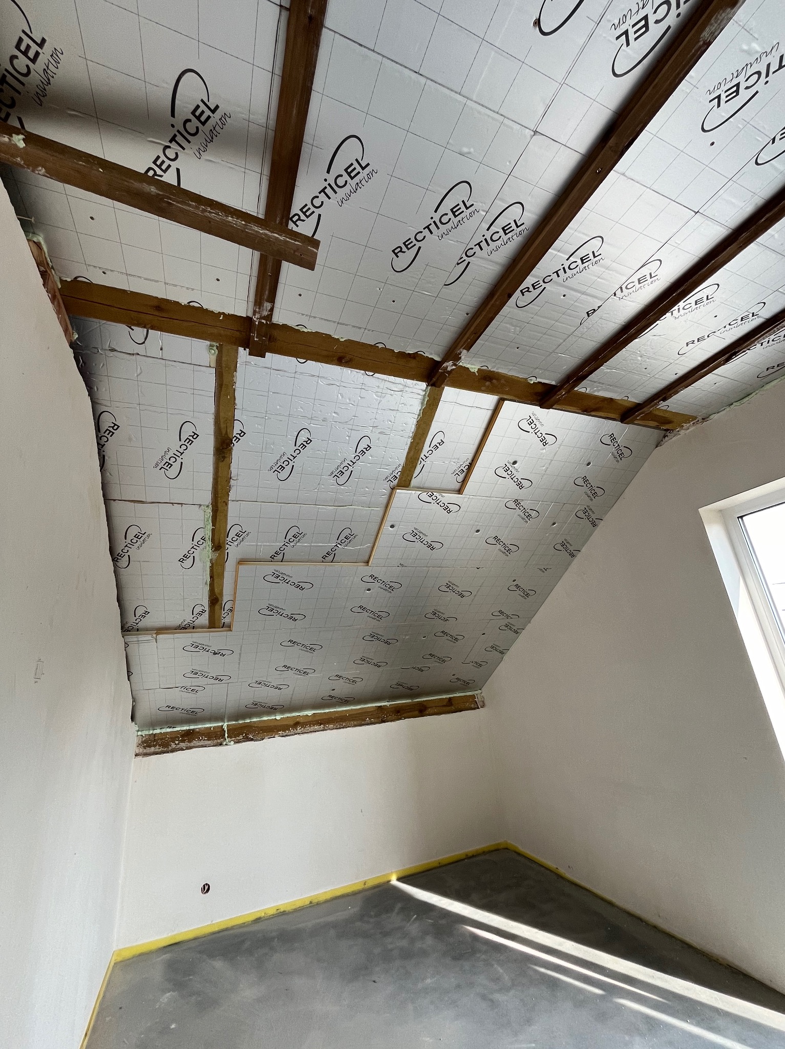 Poddasze w trakcie ocieplania, widoczne drewniane elementy konstrukcyjne dachu i płyty izolacyjne z nadrukowanym logo, szczeliny wypełnione pianką montażową.