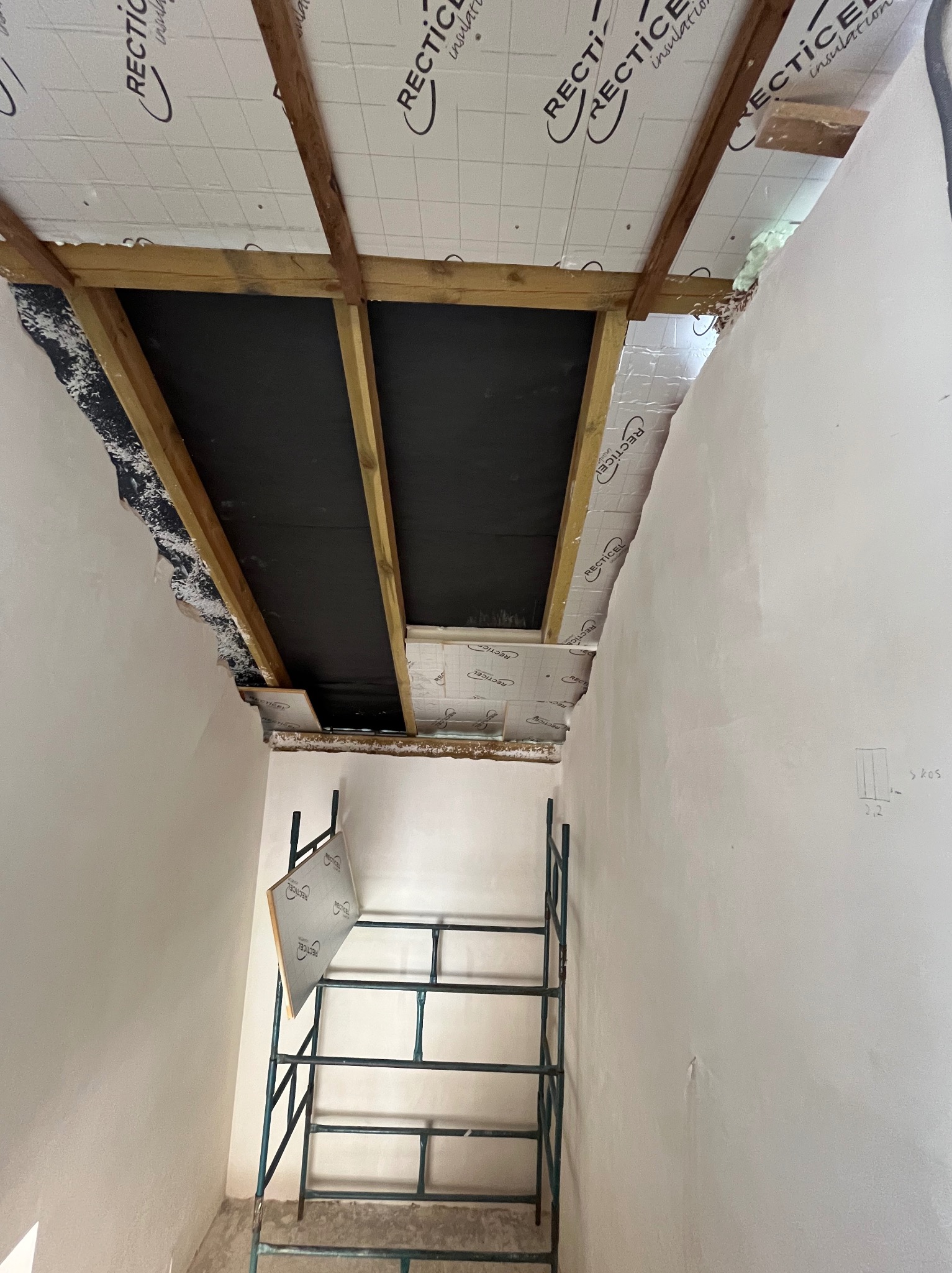 Wnętrze poddasza w trakcie ocieplania, widoczna drewniana konstrukcja dachu z izolacją termiczną Recticel, rusztowanie z fragmentem płyty izolacyjnej, surowe ściany w tle.