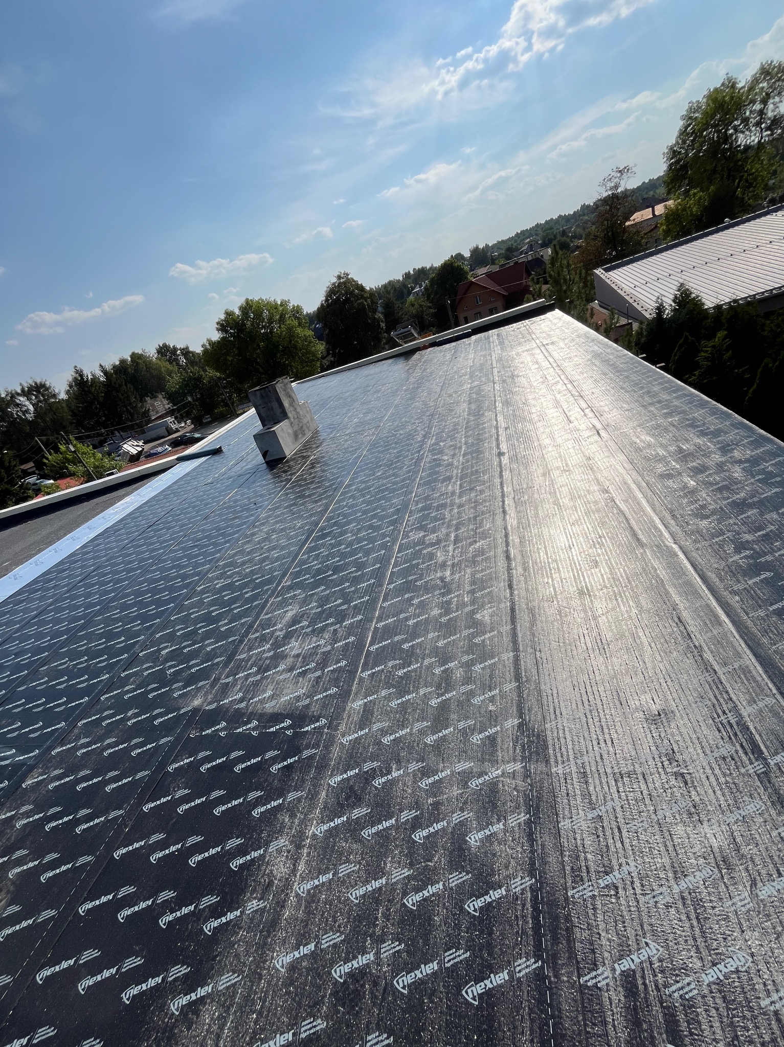 Hydroizolacja dachu płaskiego wykonana z papy termozgrzewalnej z widocznym kominem i fragmentem zabudowy w tle.