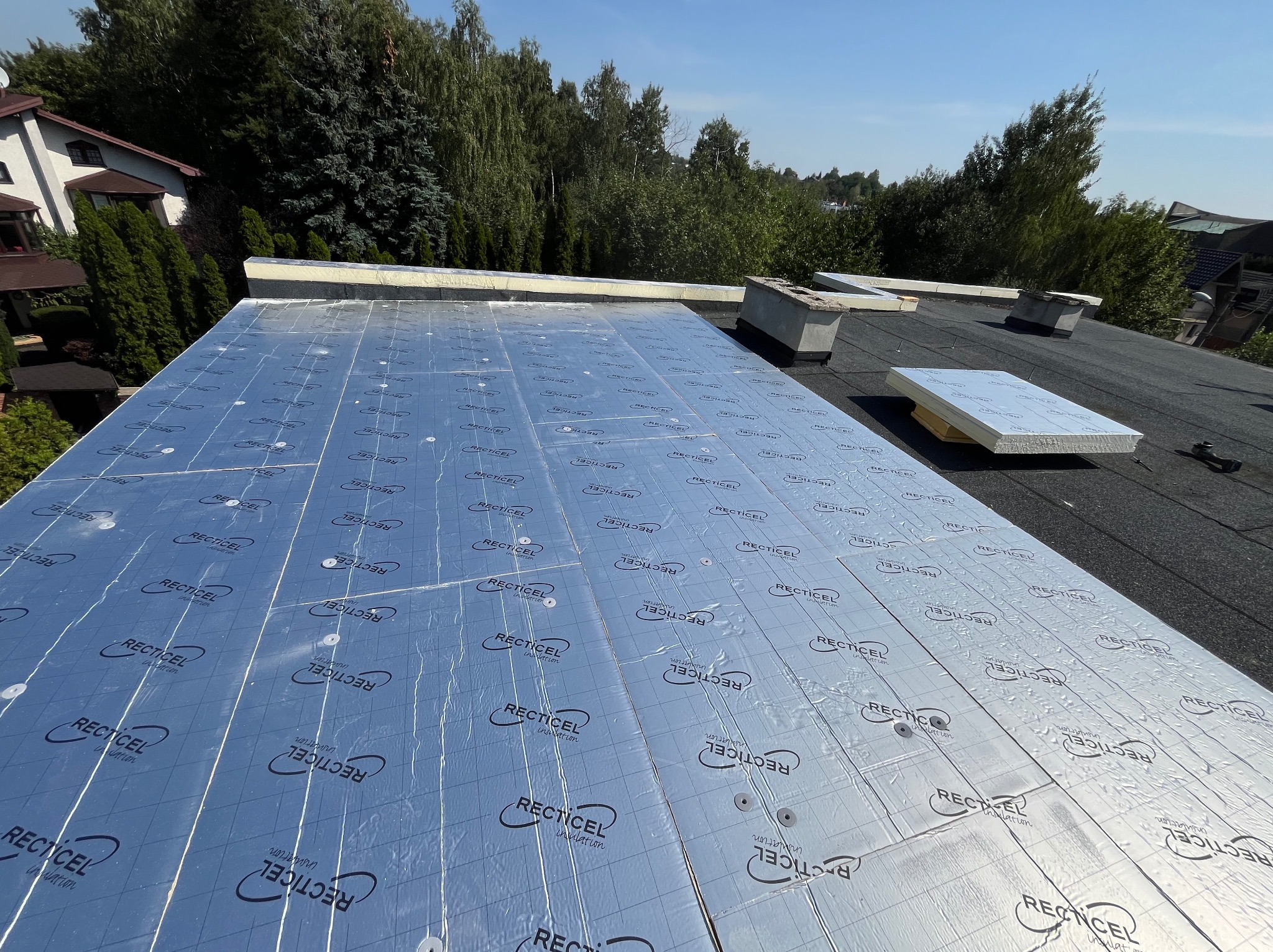 Dach pokryty membraną izolacyjną Recticel, widoczne kominy wentylacyjne i fragmenty styropianu, w tle zielone drzewa i dom