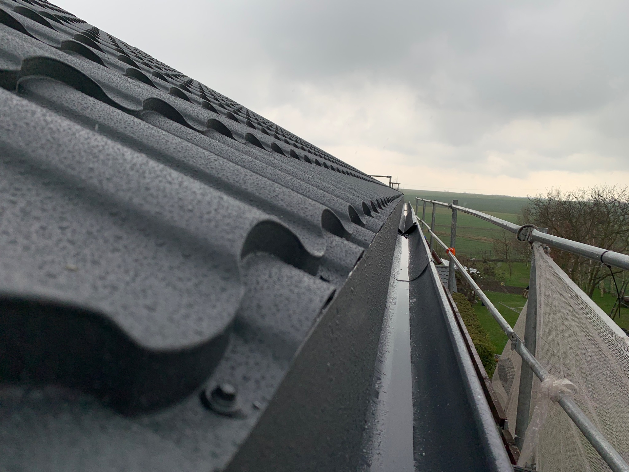 Nowy, grafitowy dach z blachodachówki modułowej, widoczny z rusztowania, z kroplami deszczu na powierzchni i fragmentem rynny wypełnionej wodą. W tle pochmurne niebo i zieleń.