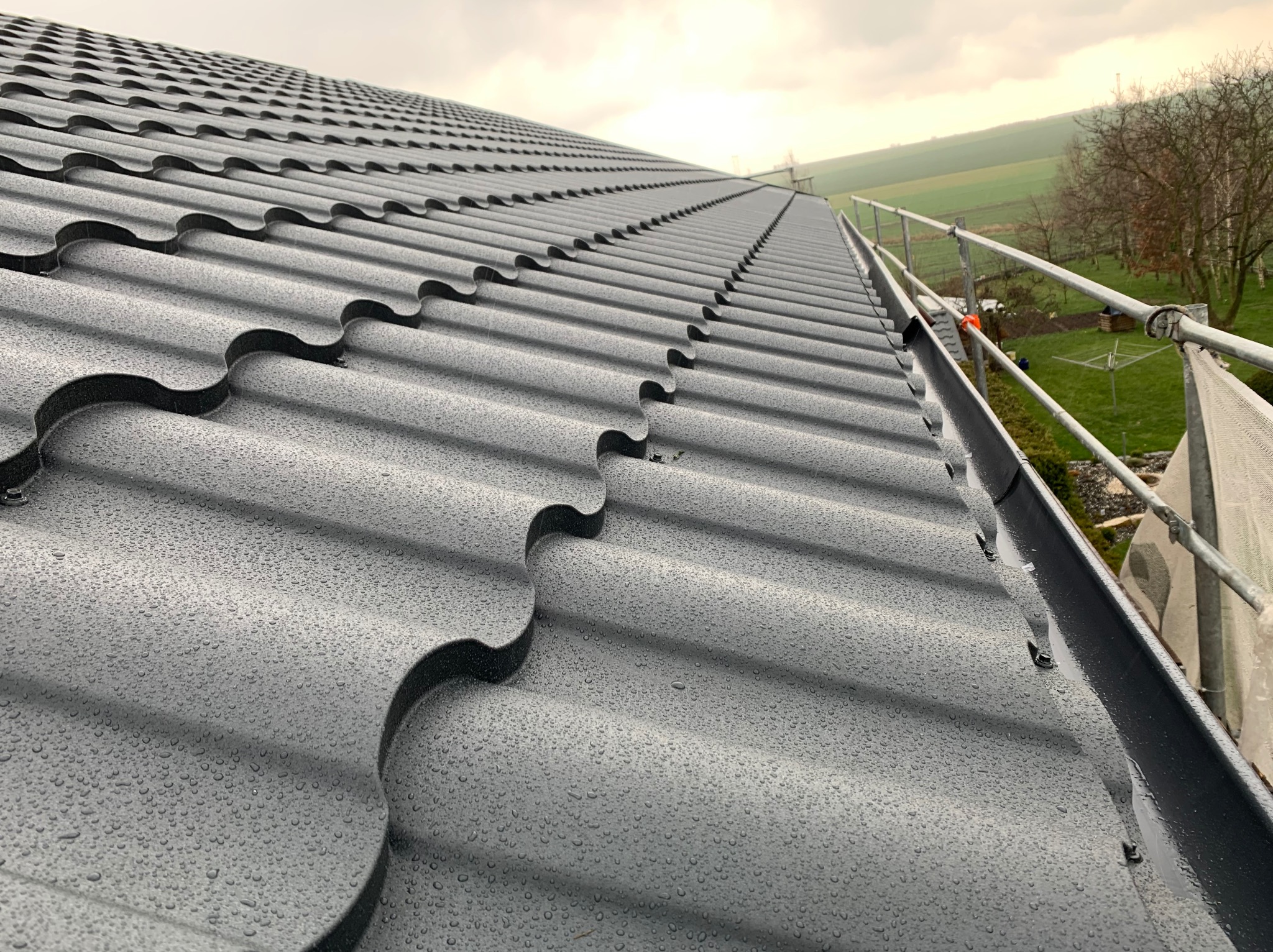 Nowy, grafitowy dach z blachodachówki modułowej po deszczu, widoczne krople wody, w tle rusztowanie i krajobraz polny.