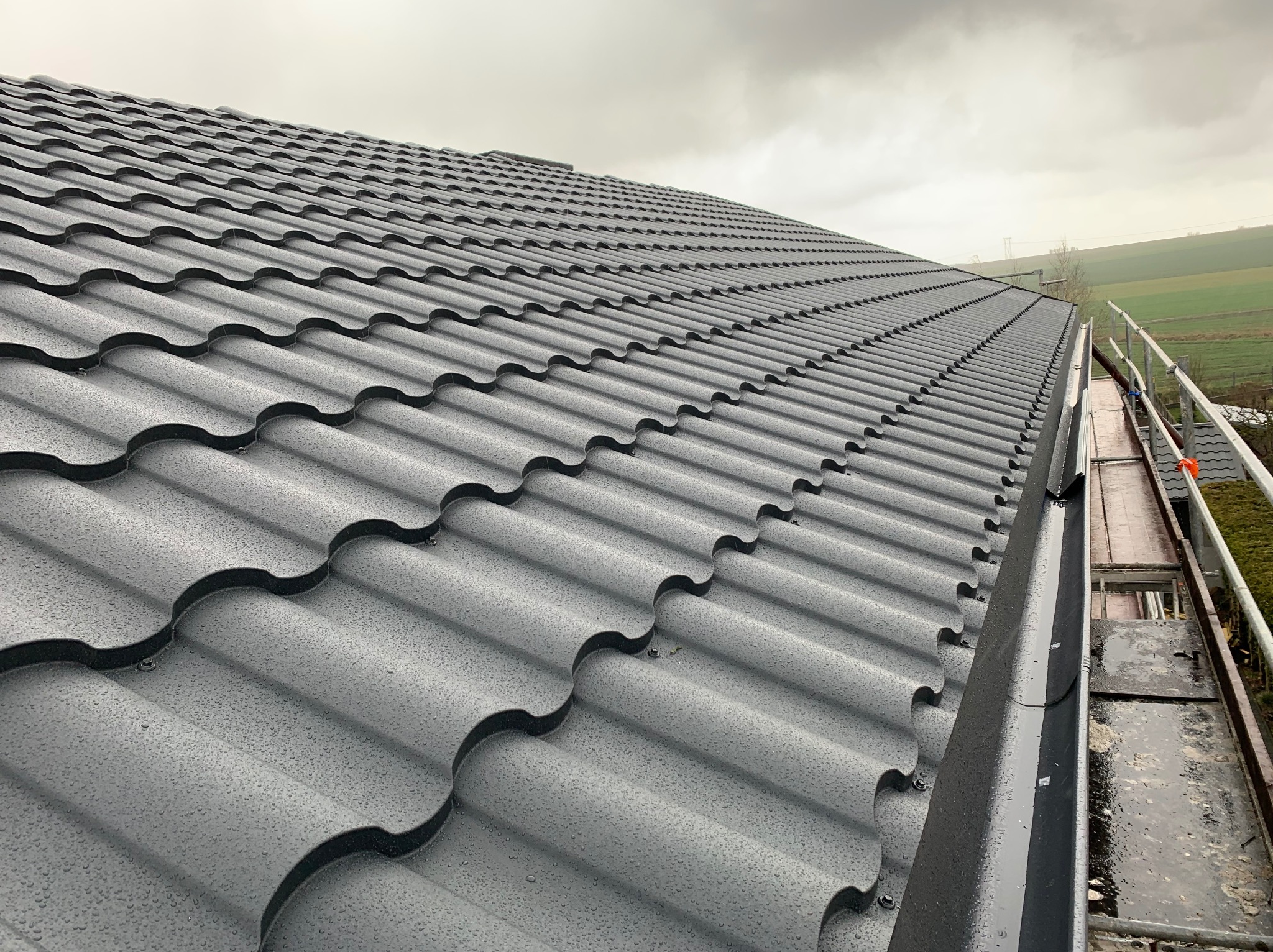 Nowy, grafitowy dach z metalowej dachówki modułowej, pokryty drobnymi kroplami deszczu, widoczny z boku, z fragmentem rynny i rusztowania.