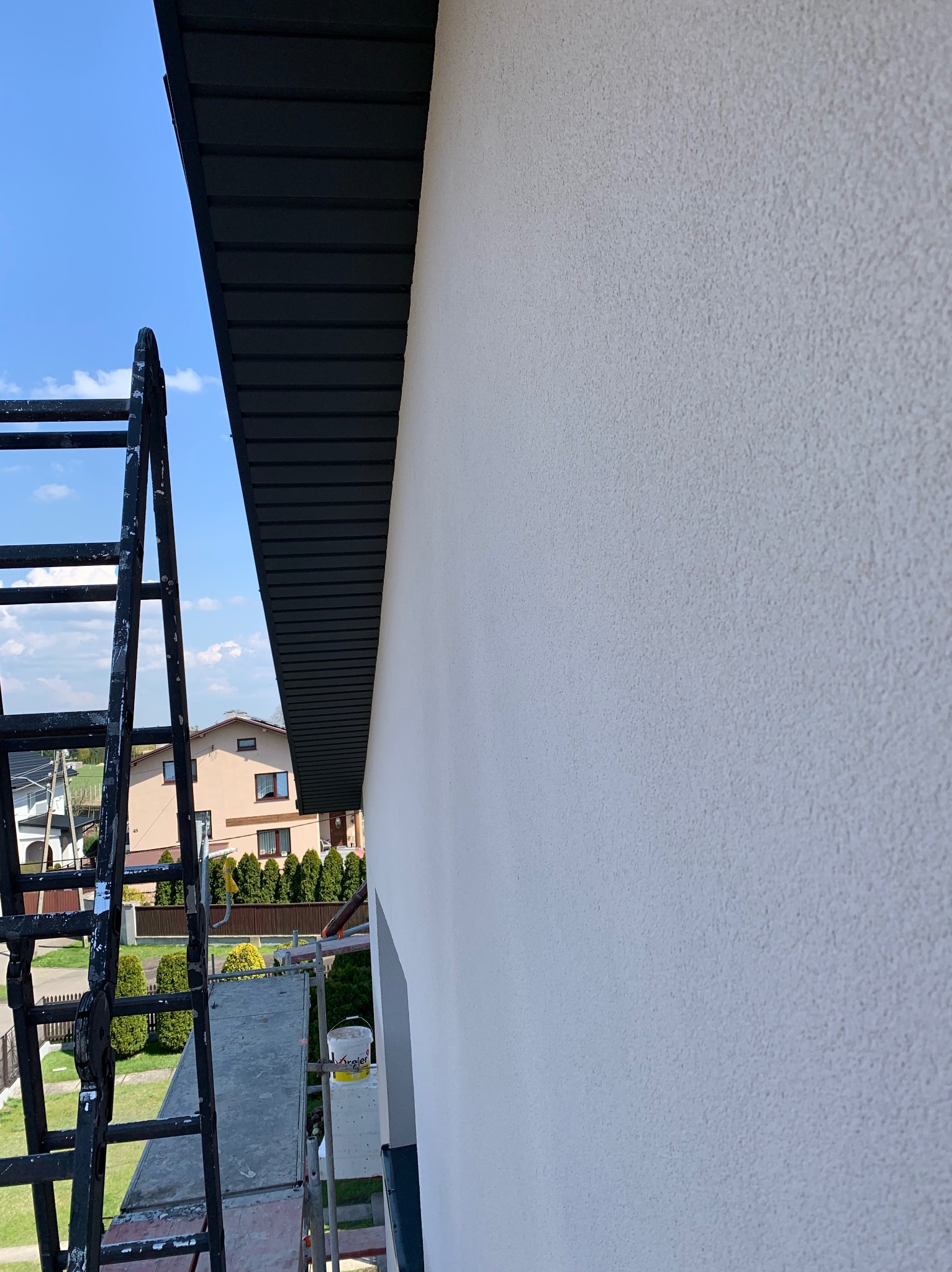 Świeżo pomalowana, jasna elewacja domu z widoczną drabina i fragmentem ciemnej podbitki dachowej na tle błękitnego nieba.