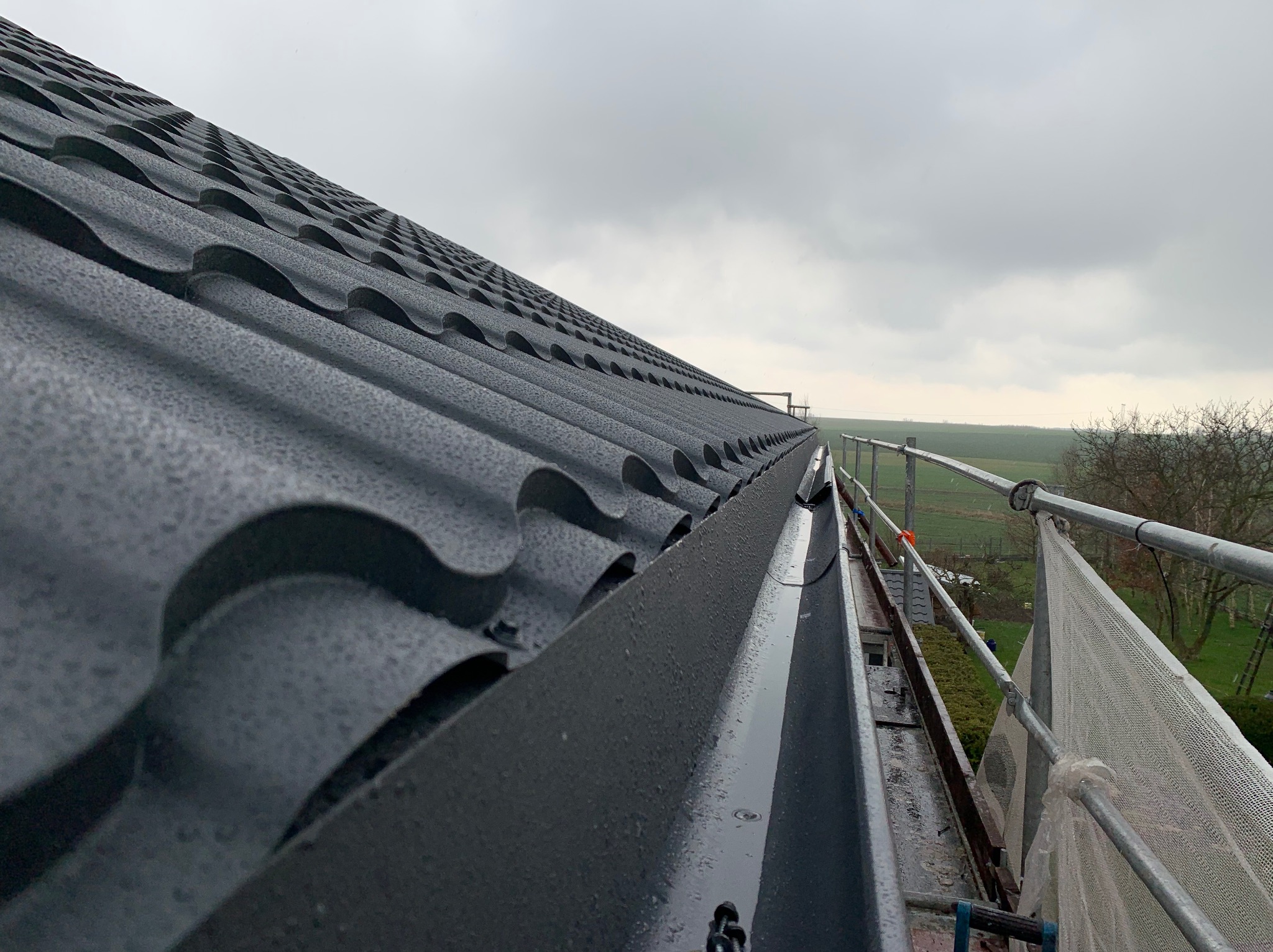 Nowy, grafitowy dach z blachodachówki modułowej, widok z rusztowania, z widoczną rynną i kroplami deszczu, na tle pochmurnego nieba i zieleni w oddali.