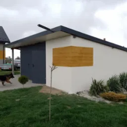 Nowoczesny garaż z elewacją w kolorze jasnej szarości z dekoracyjnym panelem imitującym drewno, ciemne grafitowe drzwi garażowe, trawnik, pies owczarek niemiecki w ruchu, pochmurne niebo.