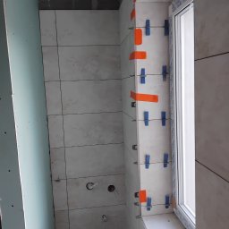Marbud - Układanie płytek w łazience, widoczne niebieskie klipsy poziomujące i pomarańczowa taśma. Ściana z płyt karton-gips po lewej, okno z parapetem po prawej.