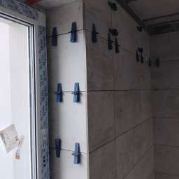 Marbud - Ściana w trakcie układania płytek ceramicznych, widoczne niebieskie klipsy poziomujące. Fragment okna z naklejkami w lewym górnym rogu. Pomarańczowe elementy mocujące.