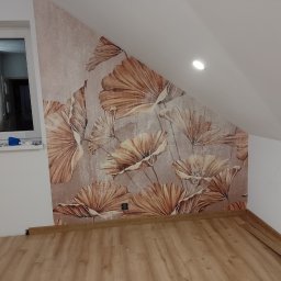 MARBUD MAREK MAJCHERCZYK - Ściana w pokoju na poddaszu z tapetą w kwiaty, drewniana podłoga. Widoczne gniazdko elektryczne i listwy przypodłogowe. Okno z trzema pionowymi lampami w tle.