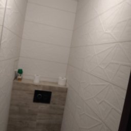 Marbud - Nowoczesna toaleta z geometrycznymi płytkami ściennymi i podtynkową spłuczką. Minimalistyczny design, jasne kolory, widoczne akcesoria toaletowe.