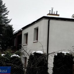 MARBUD MAREK MAJCHERCZYK - Biała elewacja domu jednorodzinnego z brązowymi ramami okiennymi, widoczna zza ogrodzenia i krzewów pokrytych śniegiem w pochmurny dzień.