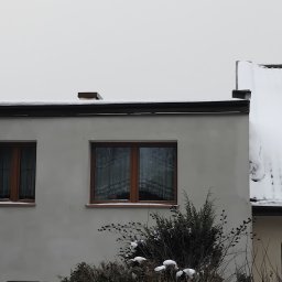 MARBUD MAREK MAJCHERCZYK - Elewacja budynku w zimowej scenerii, z widocznymi oknami z drewnianą stolarką i śniegiem na dachu. Szara kolorystyka elewacji i otoczenia.