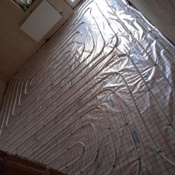 MARBUD MAREK MAJCHERCZYK - Instalacja ogrzewania podłogowego z rurkami na folii izolacyjnej, widok z góry, surowy stan wykończenia wnętrza, grzejnik na ścianie w tle.