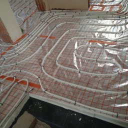 MARBUD MAREK MAJCHERCZYK - Białe rury ogrzewania podłogowego ułożone na folii z czerwoną siatką, przymocowane czarnymi klipsami. Widoczne fragmenty ścian w surowym stanie.