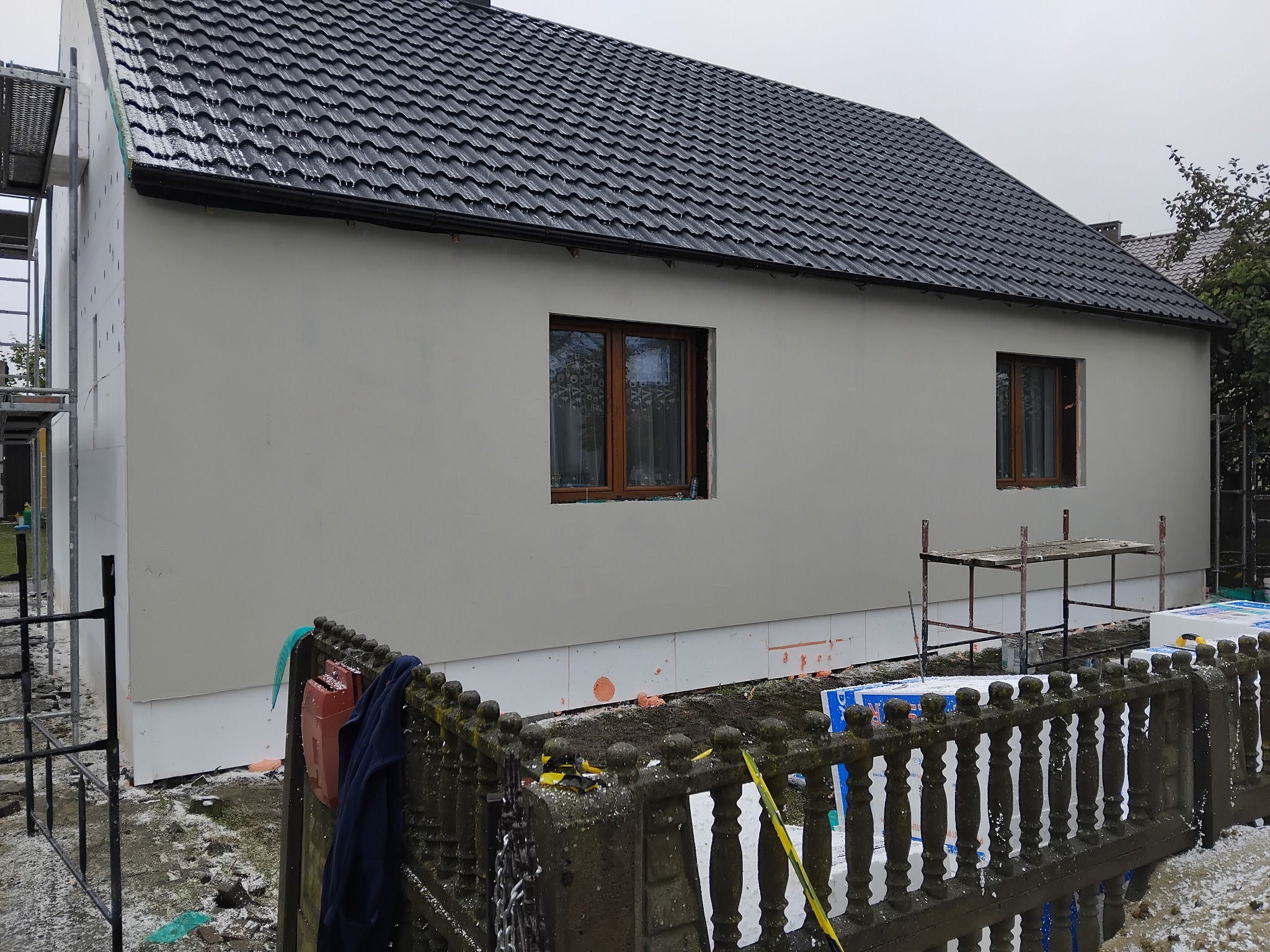 Dom jednorodzinny po nałożeniu jasnoszarego tynku elewacyjnego. Widoczne rusztowania, okna z brązowymi ramami i fragment starego, betonowego ogrodzenia.