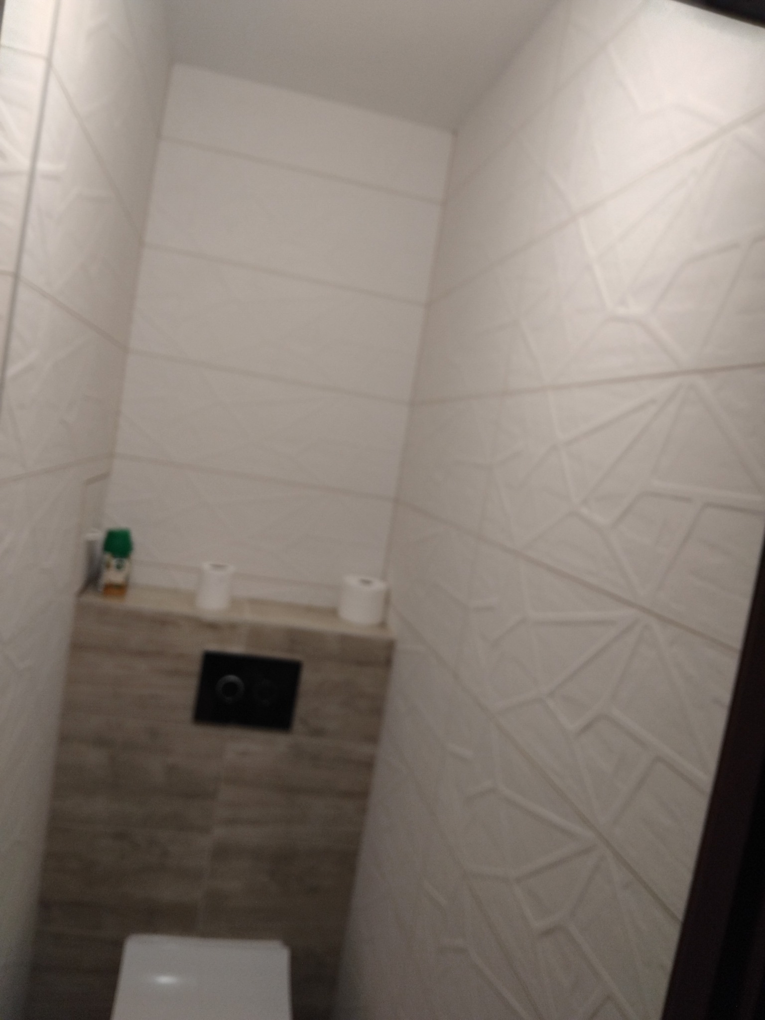 Nowoczesna toaleta z geometrycznymi płytkami ściennymi i podtynkową spłuczką. Minimalistyczny design, jasne kolory, widoczne akcesoria toaletowe.