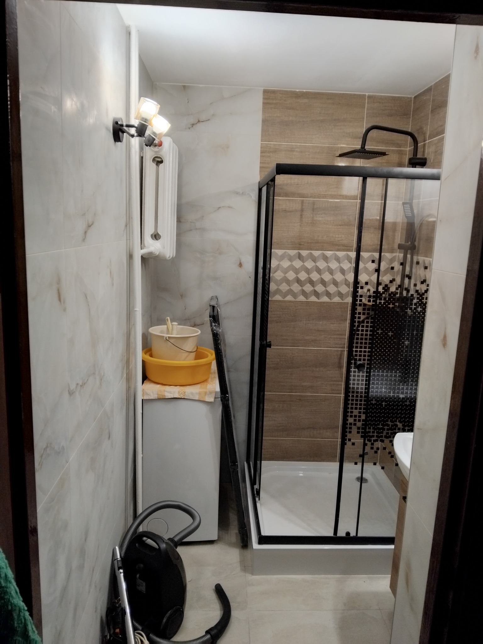 Nowoczesna łazienka po remoncie: kabina prysznicowa z czarną ramą, marmurowe płytki, grzejnik i oświetlenie. Widok z perspektywy wejścia.