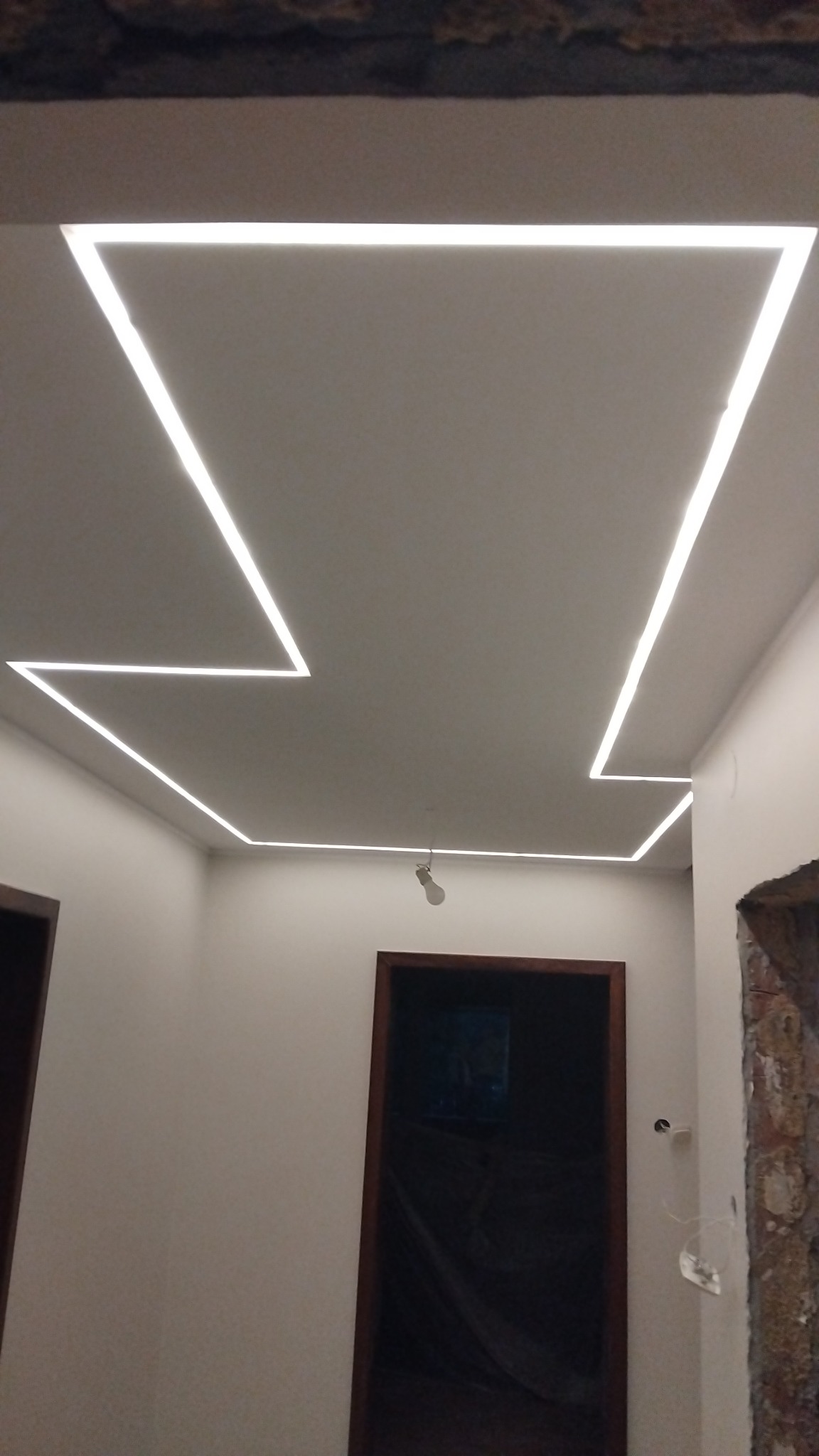 Nowoczesne oświetlenie sufitowe LED w kształcie zygzaka, w jasnym wnętrzu z widocznymi drzwiami i fragmentem ściany w trakcie prac wykończeniowych.