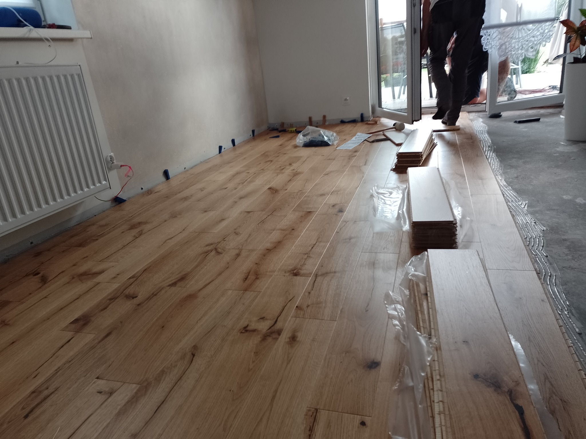 Układanie paneli podłogowych w jasnym odcieniu drewna, widoczne kliny dystansowe przy ścianie i zapas paneli w folii. Częściowo ułożona podłoga w pokoju.