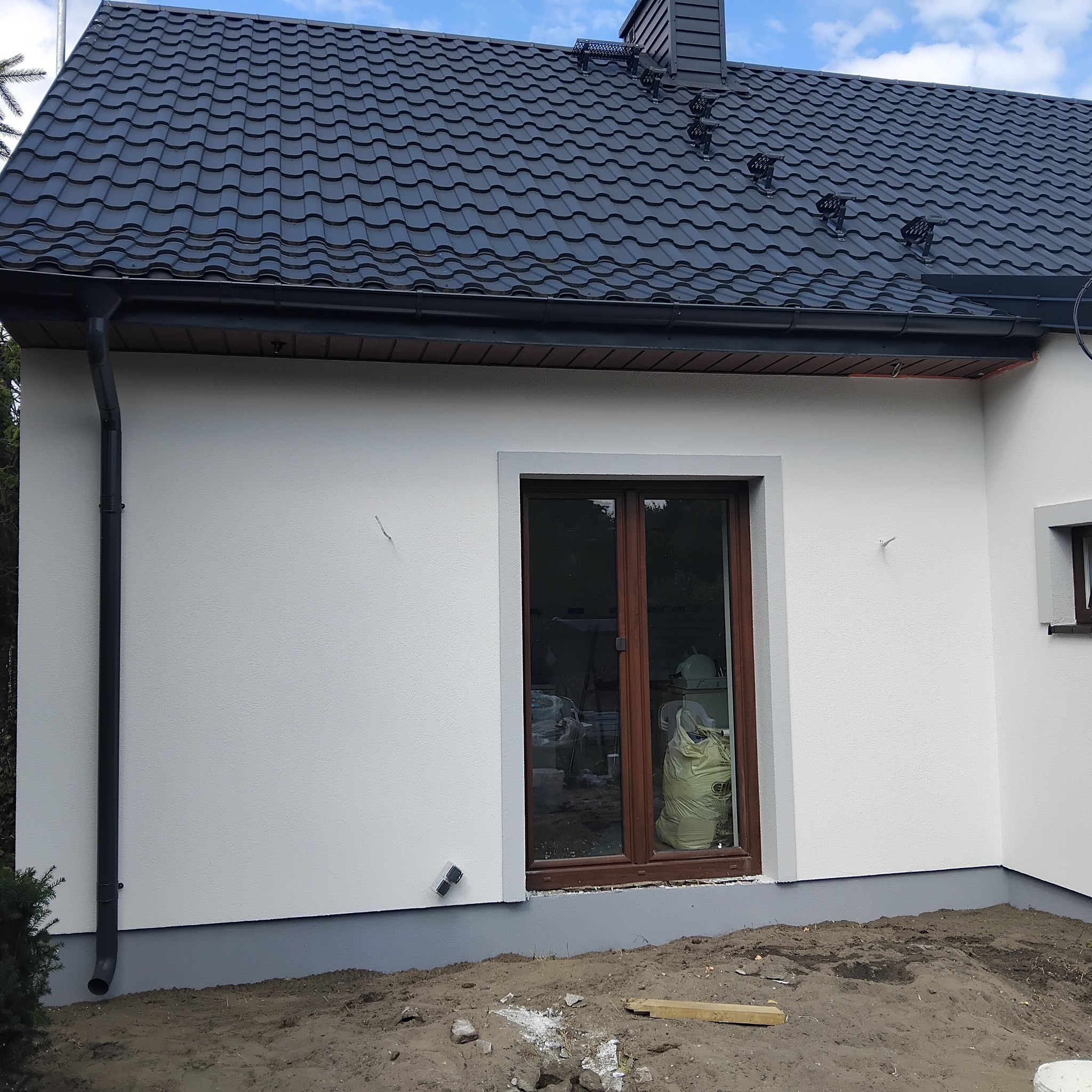 Biała elewacja domu z ciemną dachówką i brązowymi oknami. Widoczny fragment ogrodu i rynna. Świeżo położona elewacja z widocznymi detalami wykończenia.