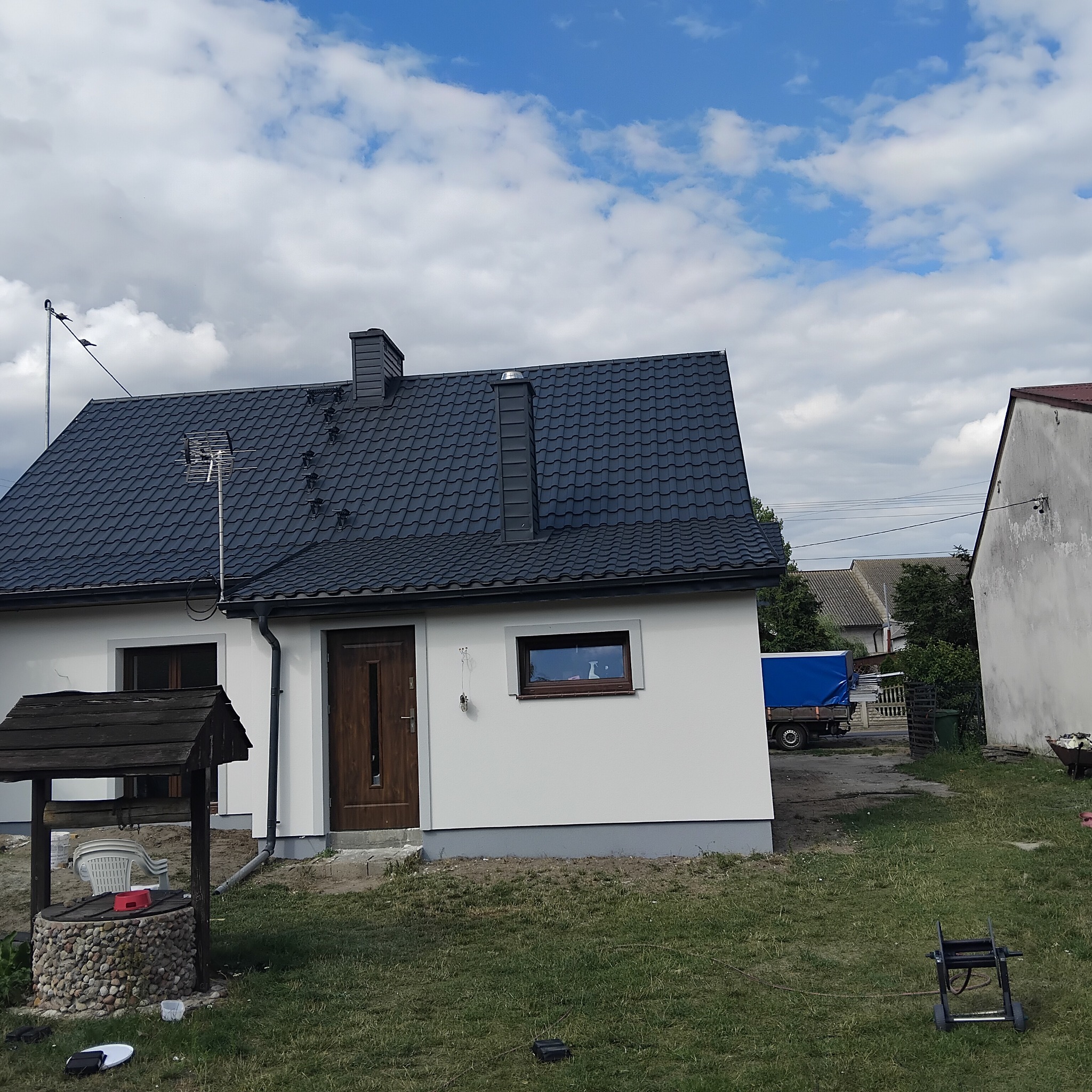 Dom jednorodzinny po remoncie elewacji z nowym dachem, studnią z kamienia na pierwszym planie i błękitnym niebem w tle. Widoczna antena na dachu.