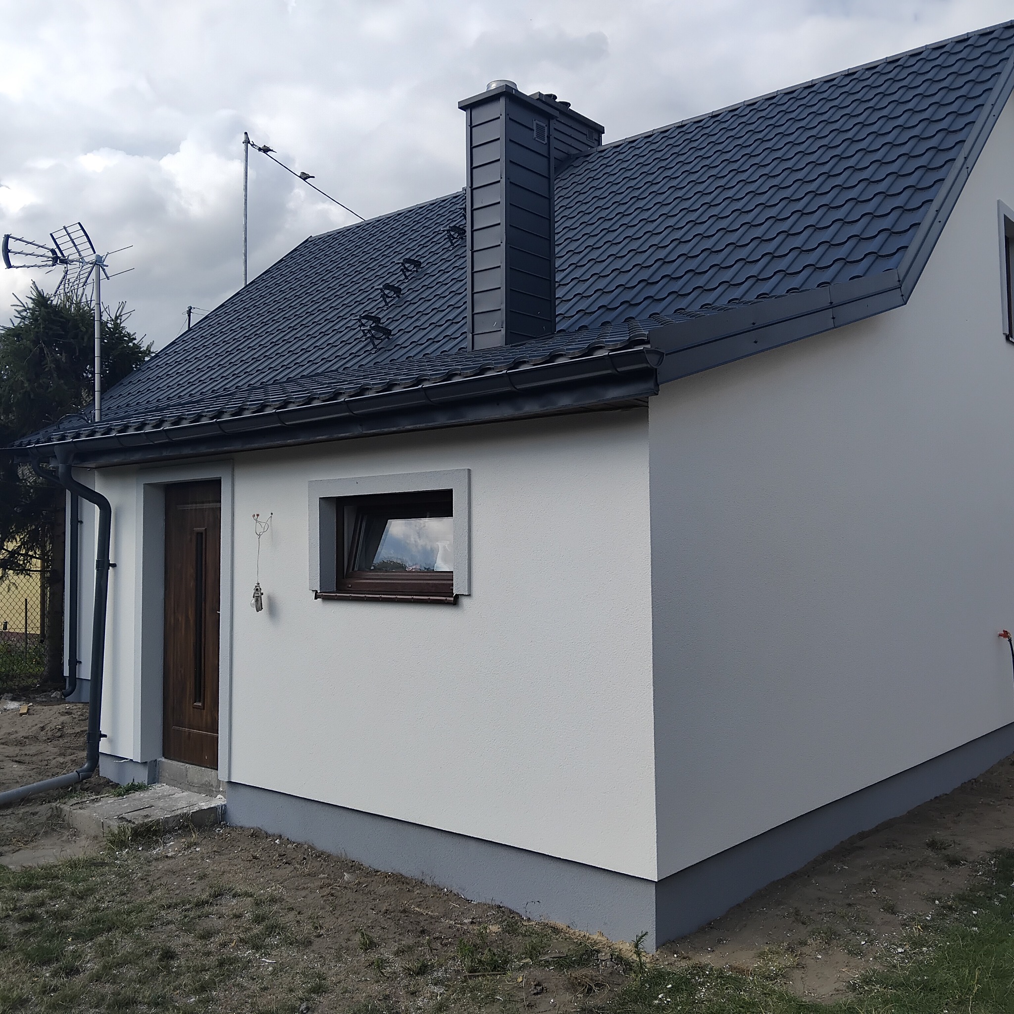 Wykończona elewacja domu jednorodzinnego w Sieradzu. Jasna fasada, ciemny dach i okna. Widoczne detale wykończeniowe, takie jak rynny i komin.