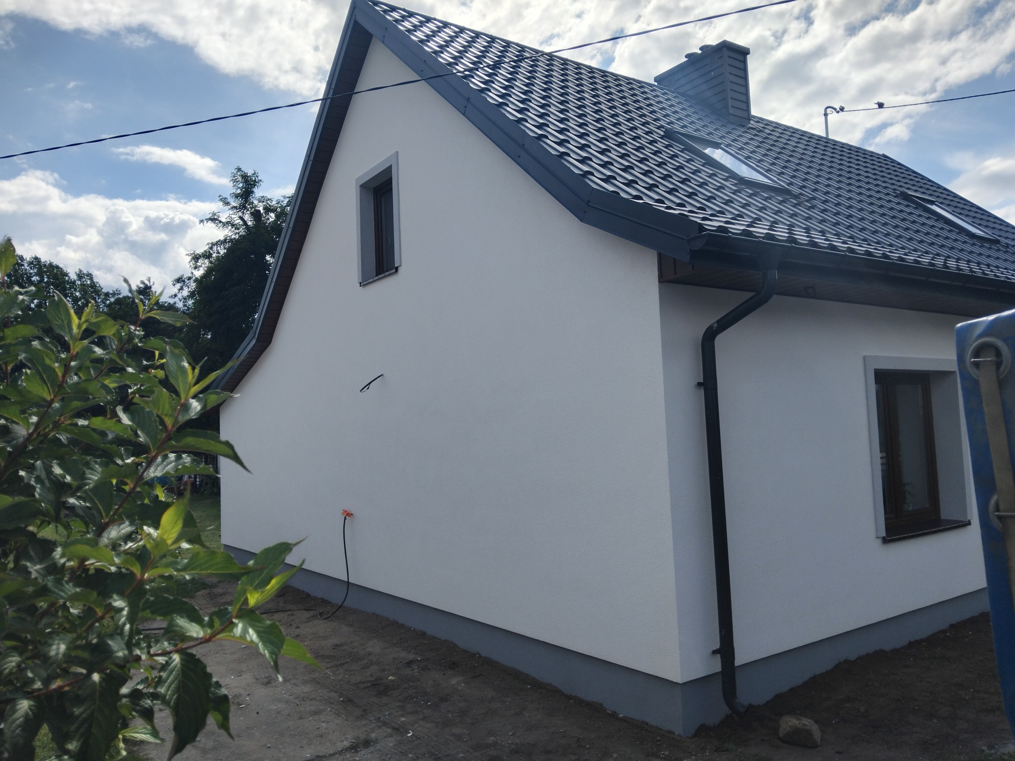 Boczny widok domu z białą elewacją i ciemnoszarą dachówką, z widocznym fragmentem zieleni po lewej stronie. Świeżo pomalowana podmurówka.