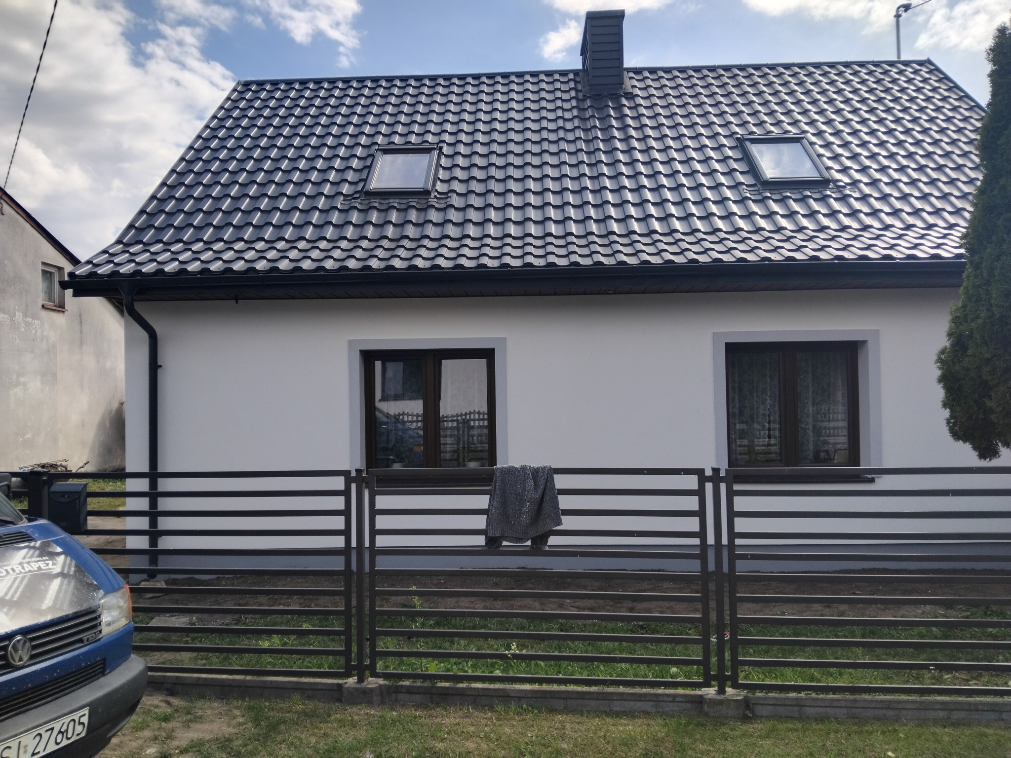Biały dom jednorodzinny z ciemną dachówką i oknami dachowymi, ogrodzony metalowym płotem. Na płocie zawieszony szary pled. Błękitny samochód częściowo widoczny po lewej.