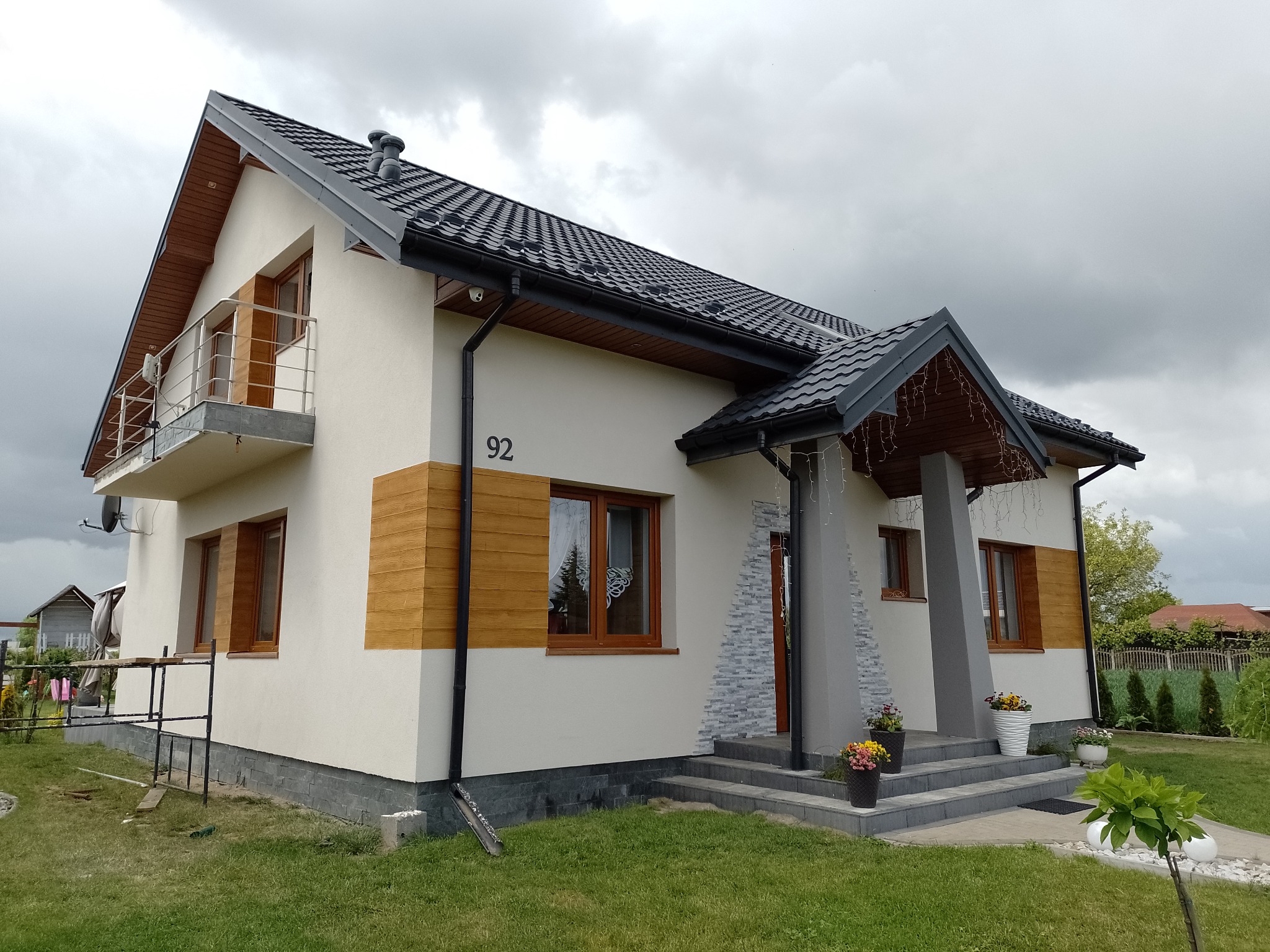 Dom jednorodzinny z balkonem, elewacją w odcieniach bieli i drewna, czarną dachówką i numerem 92. Widok z ogrodu w pochmurny dzień.