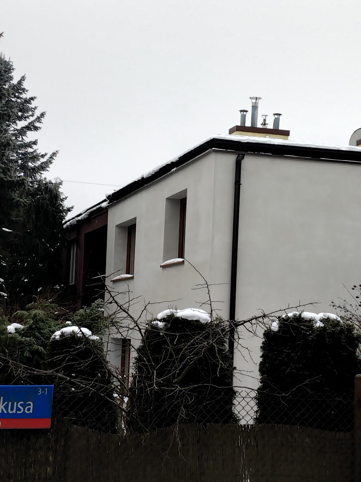 Biała elewacja domu jednorodzinnego z brązowymi ramami okiennymi, widoczna zza ogrodzenia i krzewów pokrytych śniegiem w pochmurny dzień.