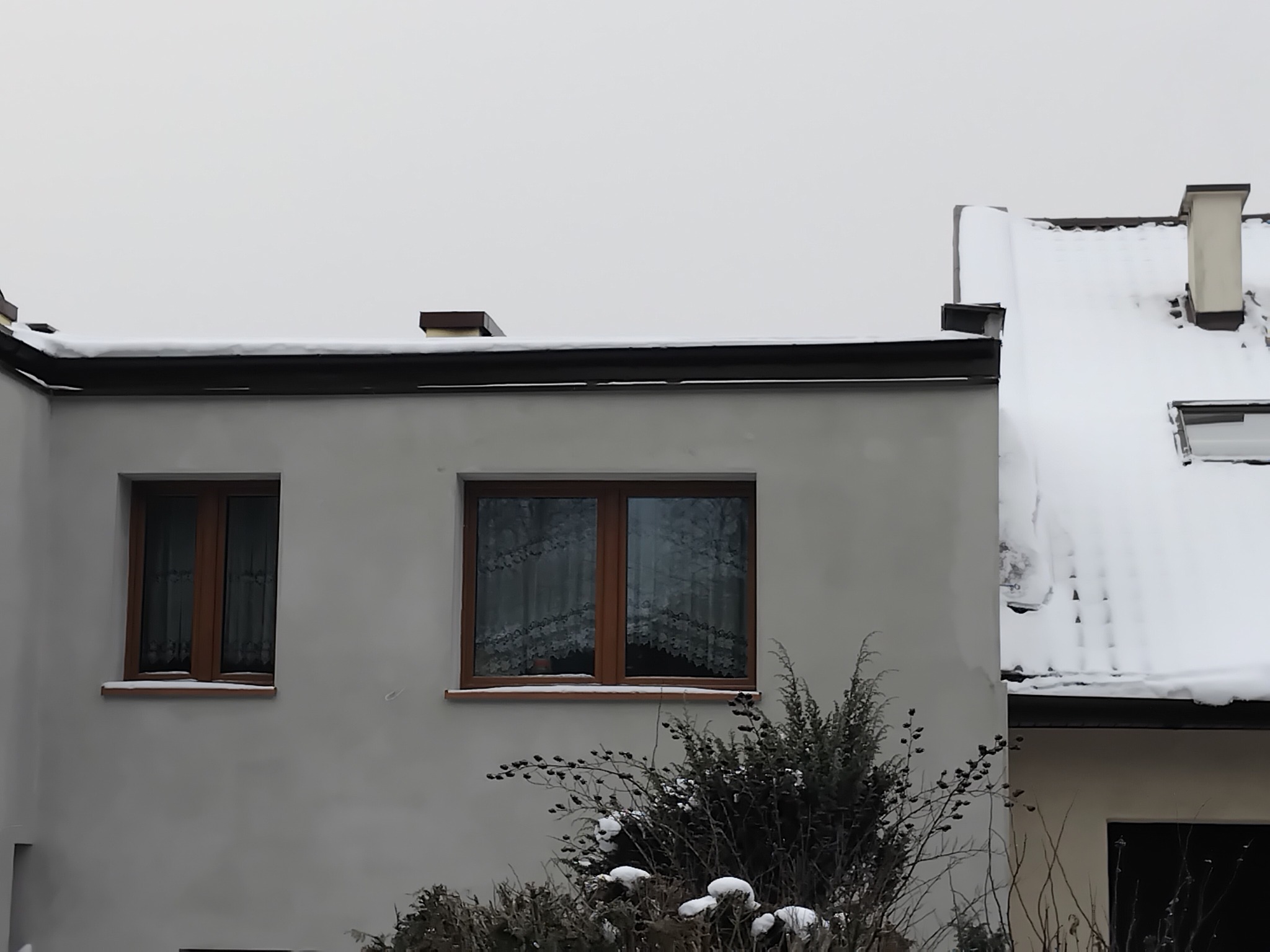 Elewacja budynku w zimowej scenerii, z widocznymi oknami z drewnianą stolarką i śniegiem na dachu. Szara kolorystyka elewacji i otoczenia.