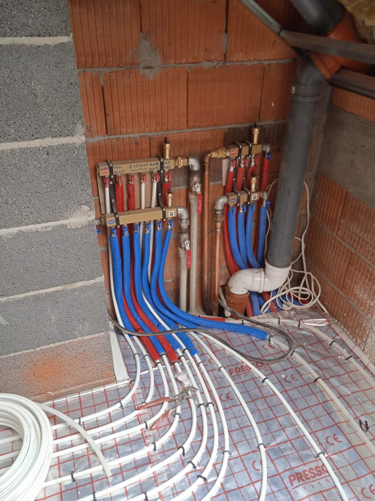 Rozdzielacz ogrzewania podłogowego z rurami w kolorach niebieskim i czerwonym, zamontowany przy ceglanej ścianie. Widoczne białe rury na folii izolacyjnej.