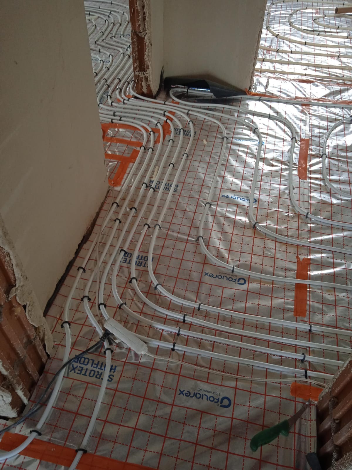 Białe rury ogrzewania podłogowego, zamocowane na folii z czerwoną siatką, widoczne fragmenty ścian w surowym stanie. Instalacja przed wylaniem posadzki.