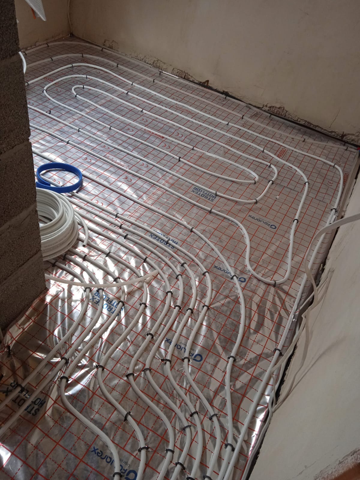 Rozłożona instalacja ogrzewania podłogowego na folii izolacyjnej z widocznymi rurami i elementami mocującymi, przygotowana do zalania wylewką.