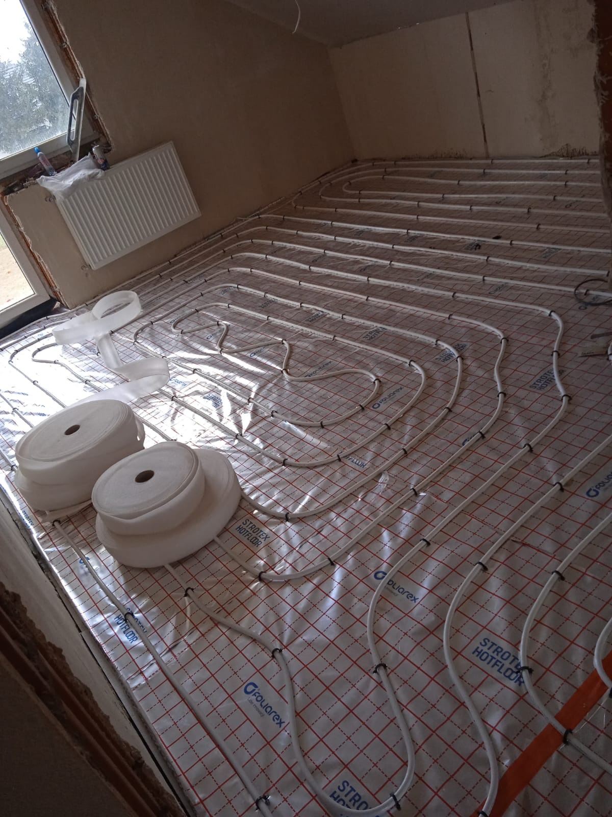 Białe rury ogrzewania podłogowego ułożone na folii z czerwoną siatką, widoczne rolki taśmy montażowej i grzejnik w tle. Etap instalacji przed wylewką.