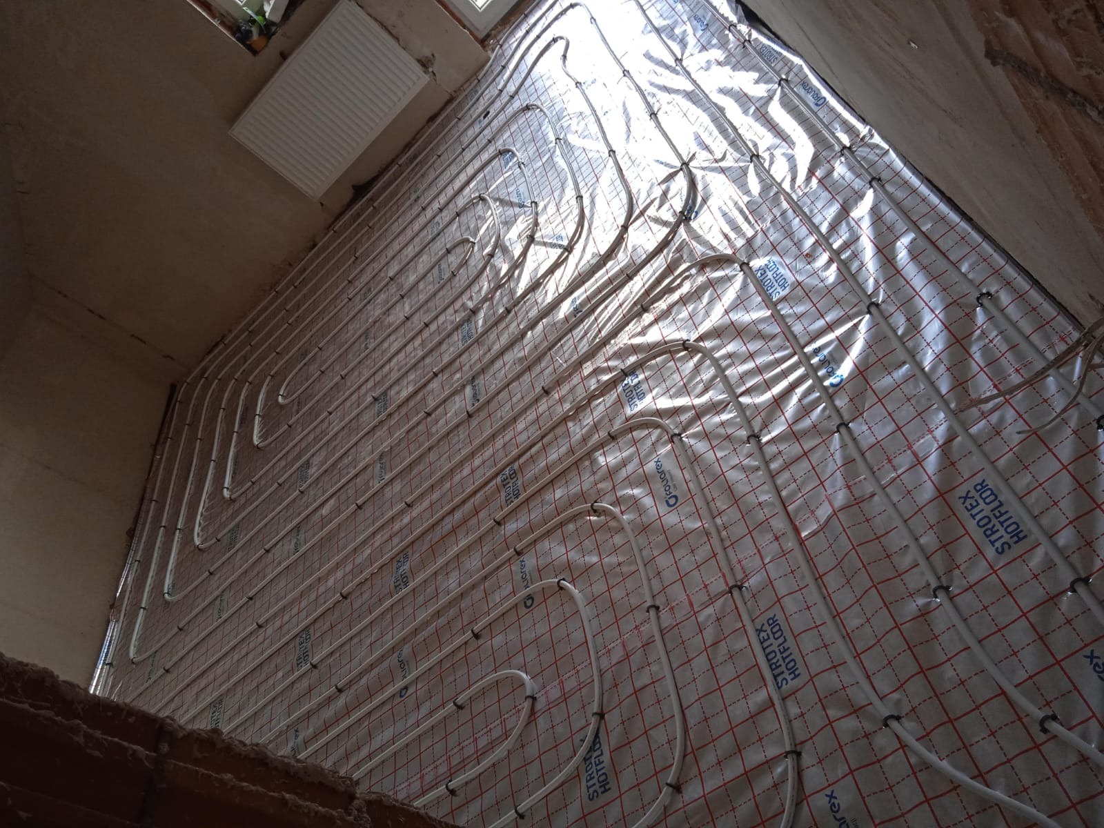 Instalacja ogrzewania podłogowego z rurkami na folii izolacyjnej, widok z góry, surowy stan wykończenia wnętrza, grzejnik na ścianie w tle.
