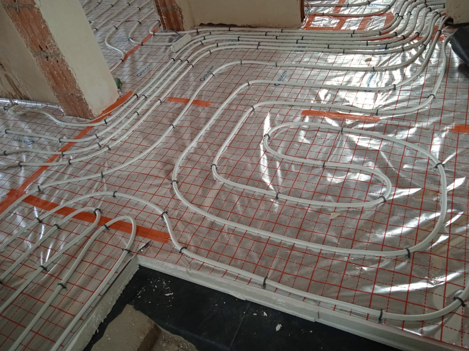 Białe rury ogrzewania podłogowego ułożone na folii z czerwoną siatką, przymocowane czarnymi klipsami. Widoczne fragmenty ścian w surowym stanie.