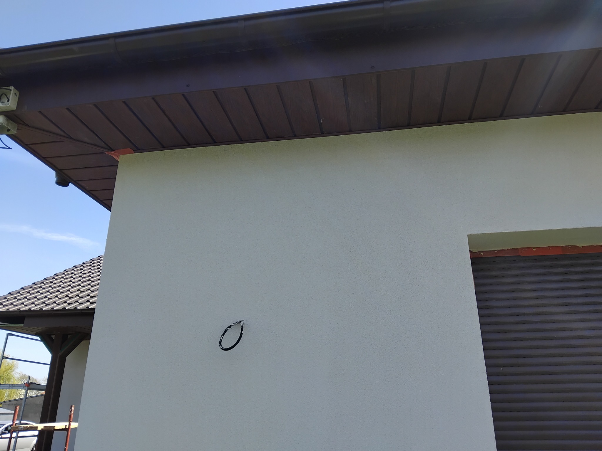 Jasna elewacja domu z brązowym podsufitką i roletą okienną. Widoczny fragment dachu i instalacja elektryczna. Świeżo położony tynk.