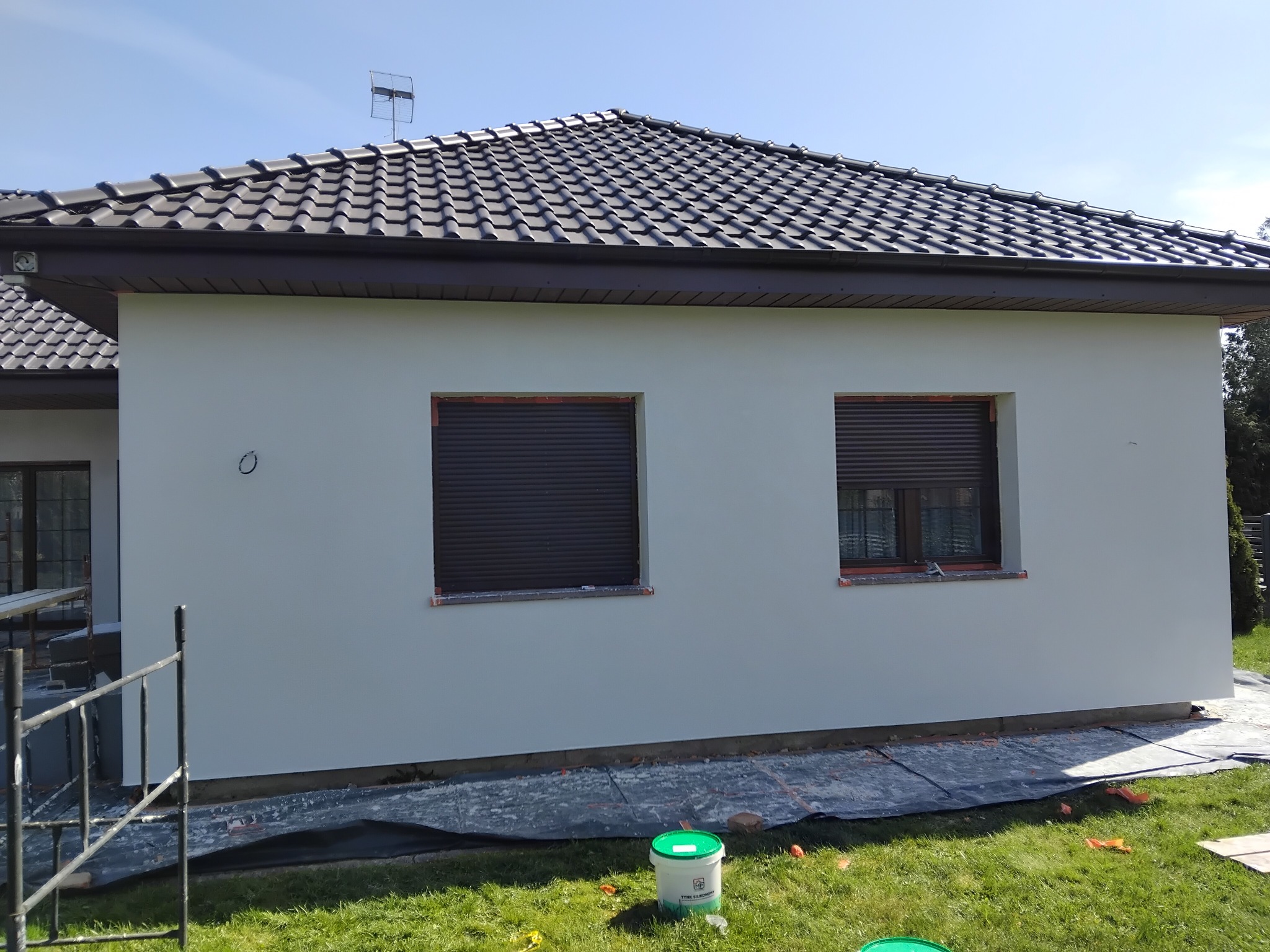 Świeżo pomalowana elewacja domu jednorodzinnego w jasnym odcieniu szarości, z widocznymi dwoma oknami z brązowymi roletami i wiaderkami farby na trawniku.