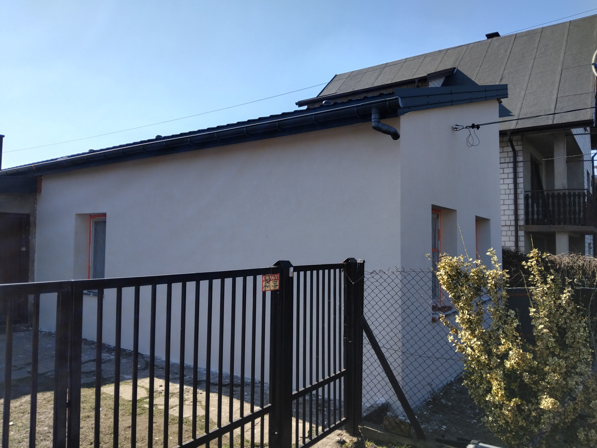 Jasna elewacja domu jednorodzinnego po remoncie, widoczna zza czarnego ogrodzenia. Świeżo otynkowana ściana, okna z pomarańczowymi ramami, błękitne niebo w tle.