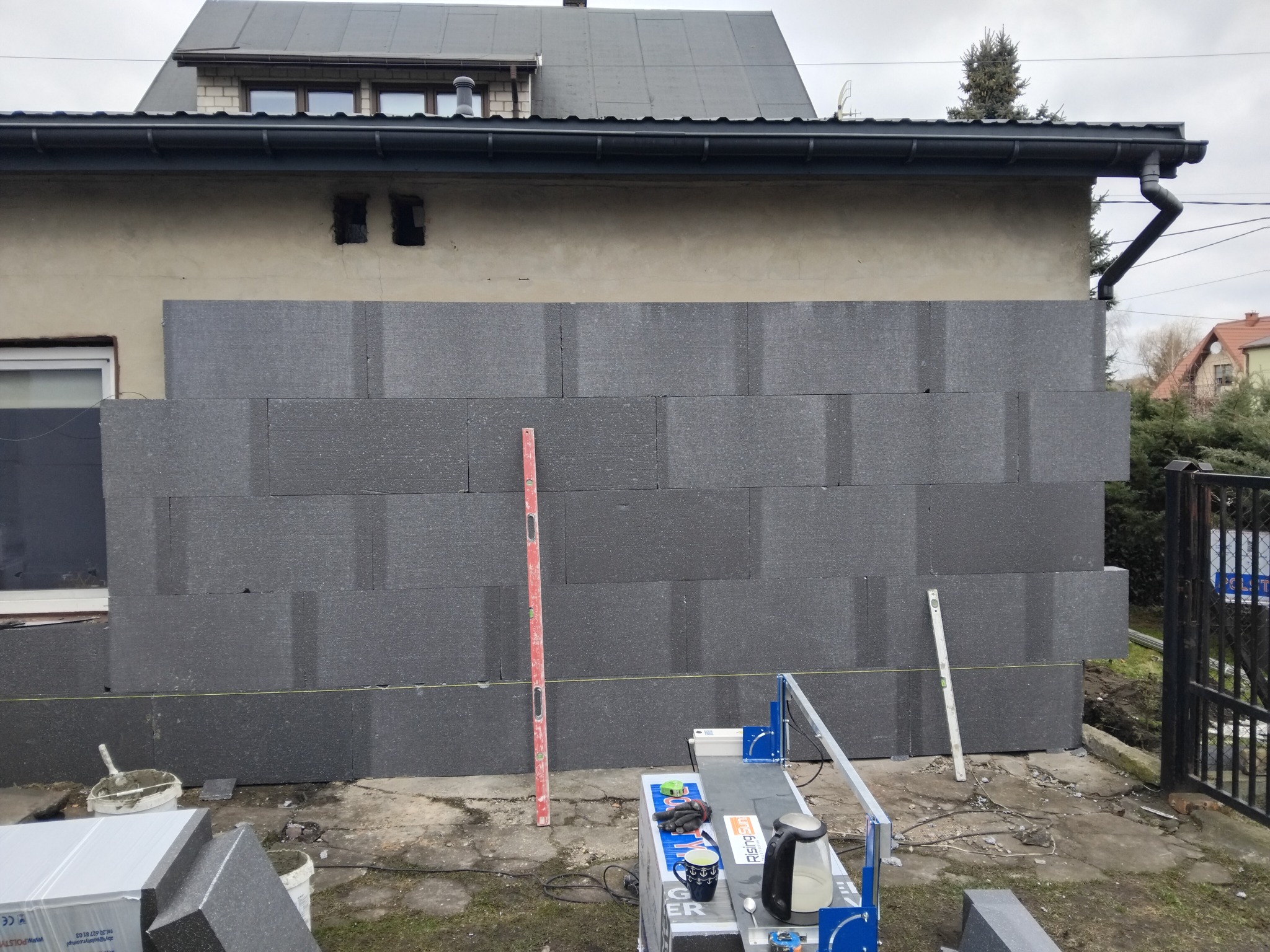 Montaż grafitowych płyt termoizolacyjnych na elewacji domu jednorodzinnego. Widoczne narzędzia pracy, poziomica i sznurek traserski. Częściowo ocieplona ściana budynku.
