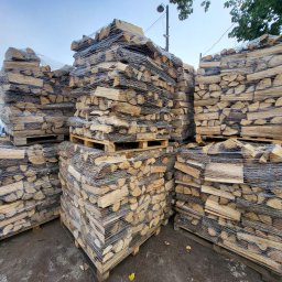 DMP Przemysław Dzięglewski - Ułożone na paletach i owinięte folią paki drewna kominkowego, widok z dołu, w tle budynek z szyldem.